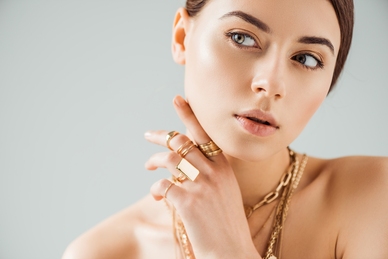 young nude woman in golden rings and necklaces looking away isolated on grey