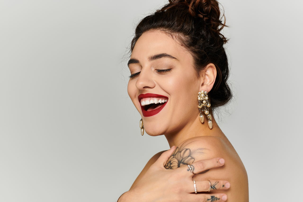Smiling young woman with a joyful expression displays her elegant earrings and tattoos.